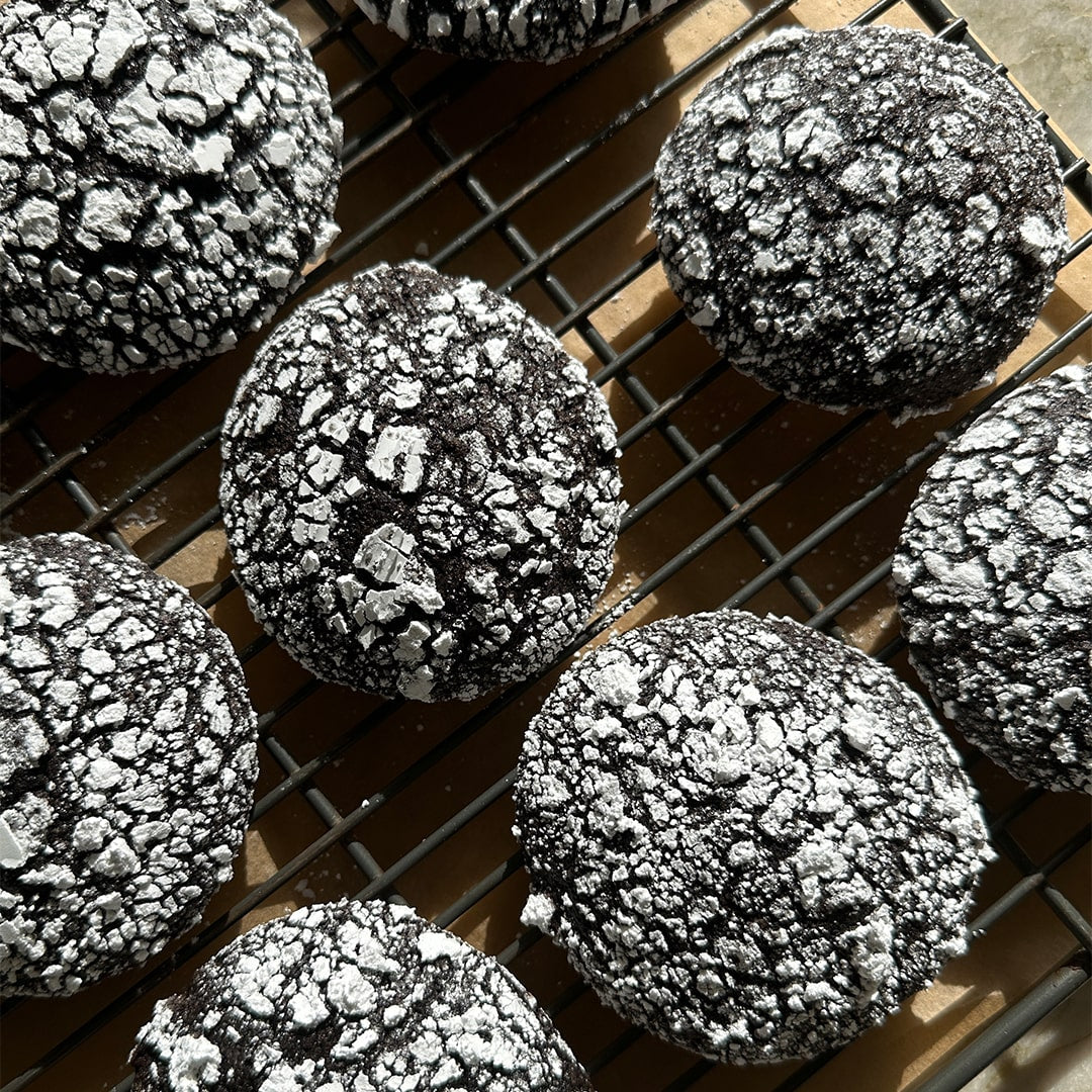Black Cocoa Crinkle Cookies (Kit) with a cracked surface and powdered sugar are cooling on a wire rack in natural sunlight.